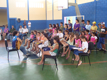 Vereadores (nas cadeiras) ao lado da diretora da Escola, Edmeia Sueli Bessa Monteiro, durante apresentação musical por Anamaria Brandi e alunos